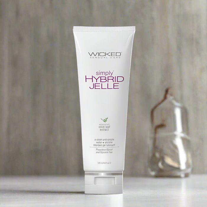 A white tube of Wicked Simply Hybrid Jelle Lubricant 4 oz by Wicked Lubes sits against a white background. The tube features purple and black text along with a green leaf design, indicating it contains olive leaf extract with moisturizing properties. Latex condom compatible, the tube holds 120 ml or 4 fl oz.