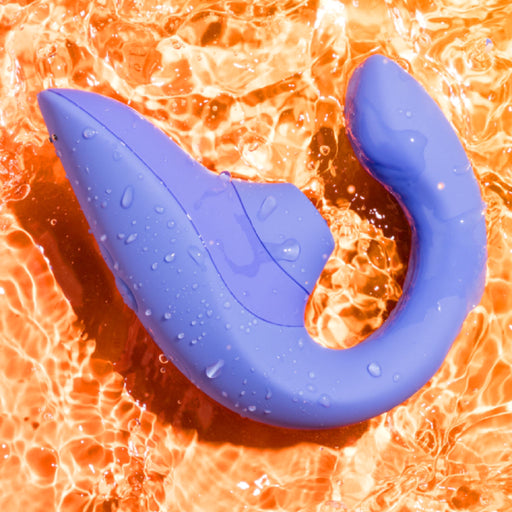 A blue Womanizer Blend Pleasure Air Clitoral & G-Spot Rabbit, designed with an ergonomic and modern aesthetic, rests on a vibrant, orange liquid background. Its sleek and curved shape suggests dual stimulation capabilities for an enhanced experience.