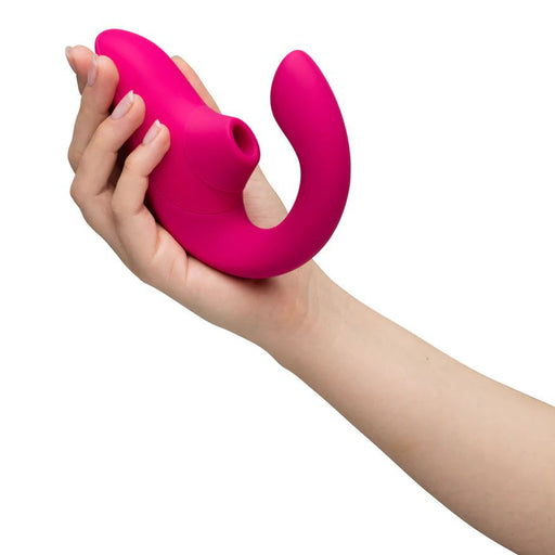 A person's hand holding the Womanizer Blend Pleasure Air Clitoral & G-Spot Rabbit in pink, a silicone vibrator with dual ends—one featuring a curved arm for G-spot stimulation and the other a rounded suction-like attachment for clitoral pleasure. The background is plain white.