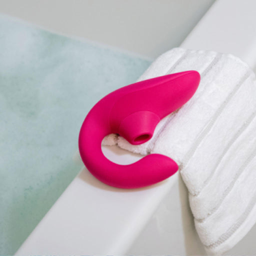 A Womanizer Blend Pleasure Air Clitoral & G-Spot Rabbit in pink rests on a white towel at the edge of a bathtub filled with light-blue water.
