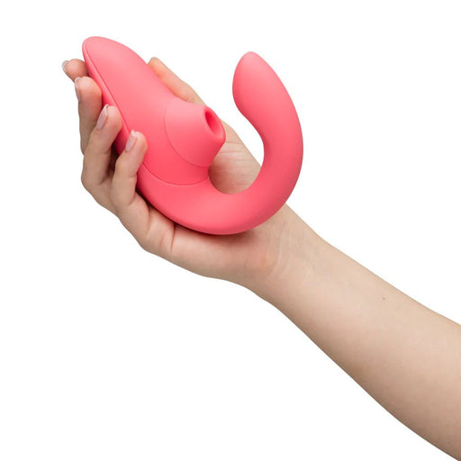 A hand holding a sleek, rose silicone Womanizer Blend Pleasure Air Clitoral & G-Spot Rabbit vibrator designed for dual stimulation, featuring a curved end and a suction feature reminiscent of the iconic Womanizer brand. The ergonomic device is modern and intended for personal use.