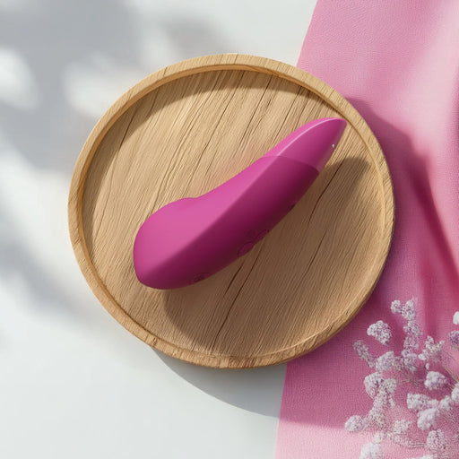 A Womanizer Enhance Pleasure Air + Vibration Clit Stimulator in pink, curvy form sits on a round wooden tray with a pink cloth and white flowers nearby, all bathed in soft natural light.