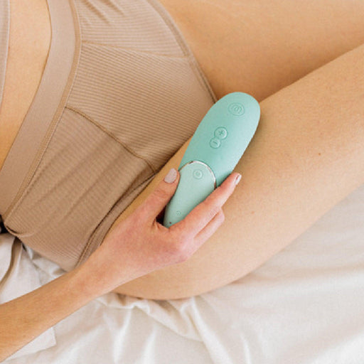 A person wearing beige underwear holds a Womanizer Next Pleasure Air Clitoral Vibrator in Sage Green against their thigh while sitting on a white bed. The device, featuring Climax Control and Pleasure Air Technology, has buttons and an indicator light. The person's nails are painted a pastel color.