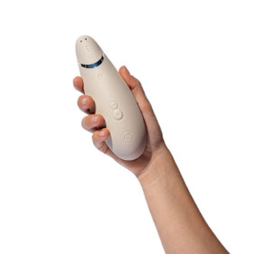 A hand holding a warm gray handheld device with a sleek, curved design, resembling the Womanizer Premium 2 Pleasure Air Clitoral Stimulator. The device features a few buttons in the middle and a small blue light indicator near the top, highlighting its advanced air pulsation stimulator technology. The background is white.