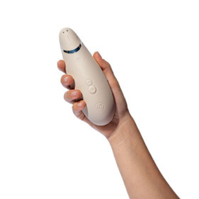 A hand holding a warm gray handheld device with a sleek, curved design, resembling the Womanizer Premium 2 Pleasure Air Clitoral Stimulator. The device features a few buttons in the middle and a small blue light indicator near the top, highlighting its advanced air pulsation stimulator technology. The background is white.