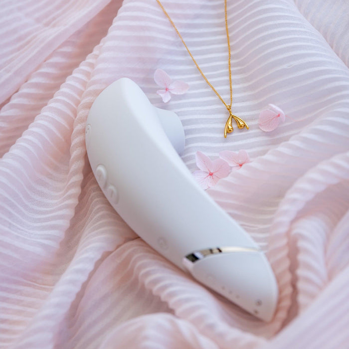 A sleek Warm Gray Womanizer Premium 2 Pleasure Air Clitoral Stimulator lies on a soft pink, pleated fabric. Next to it is a delicate gold necklace with a wishbone pendant and some small pink flower petals, creating a calm and elegant scene. The air pulsation stimulator adds an element of sophistication to the serene setting.
