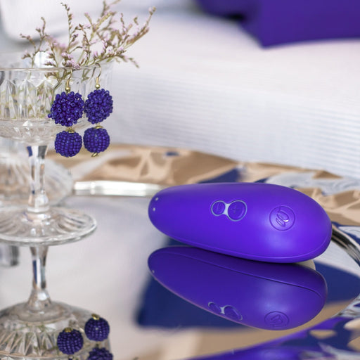 The Womanizer Starlet 3 Pleasure Air Clitoral Stimulator in Indigo, an elegant egg-shaped electronic device designed for clitoral stimulation, rests on a reflective surface beside a glass vase filled with flowers and a pair of raspberry-like beaded earrings. In the background, a softly blurred sofa and pillow contribute to the cozy ambiance of the scene.