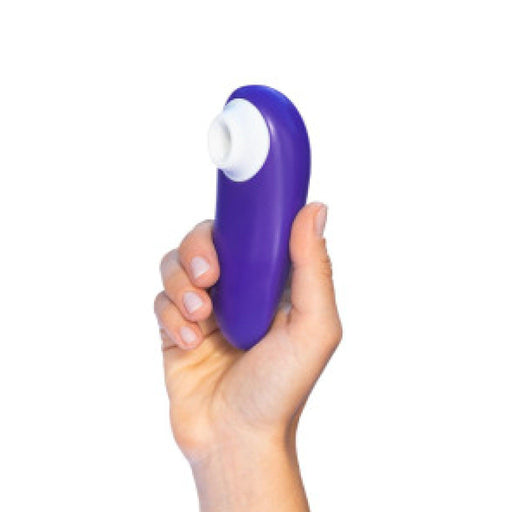 A hand holding the Womanizer Starlet 3 Pleasure Air Clitoral Stimulator in Indigo, a small purple handheld device by Womanizer with a white circular attachment at one end, against a plain white background.