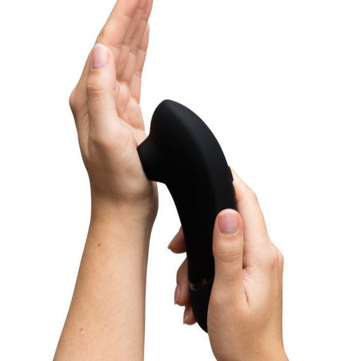 A person holds the Womanizer x We-Vibe Lust & Luxury Collection, a black, curved device, in one hand and presses it against the palm of their other hand against a white background.