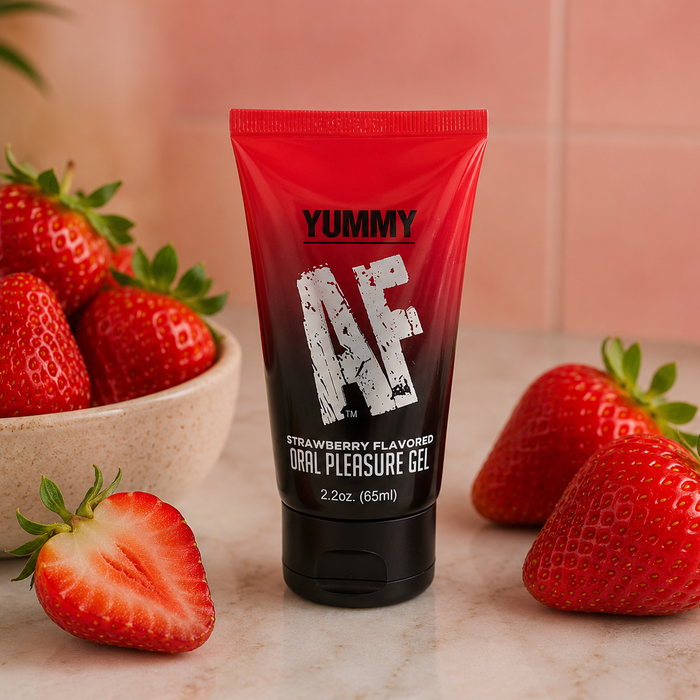 A red and black tube of Yummy AF Oral Pleasure Gel – Strawberry, a water-based oral lubricant, stands among fresh strawberries on a light surface with a pink tiled background.