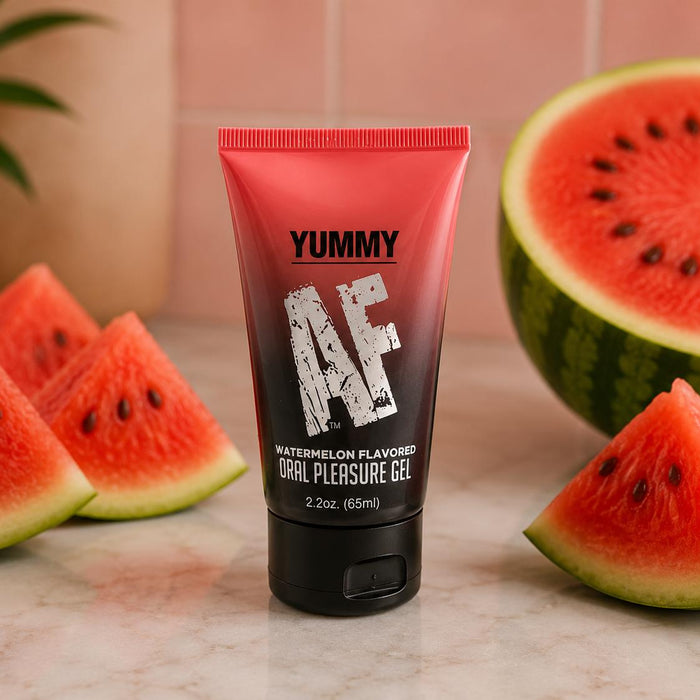 A red and black tube of Yummy AF Oral Pleasure Gel – Strawberry or Watermelon 2.2 oz sits on marble, surrounded by watermelon slices, showcasing this flavored water-based lubricant for enhanced oral pleasure.