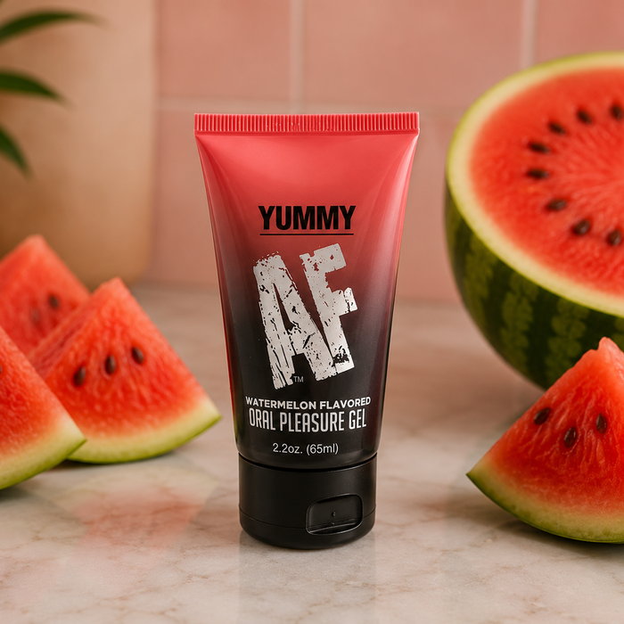 A red and black tube of Yummy AF Oral Pleasure Gel – Strawberry or Watermelon 2.2 oz sits on marble, surrounded by watermelon slices, showcasing this flavored water-based lubricant for enhanced oral pleasure.