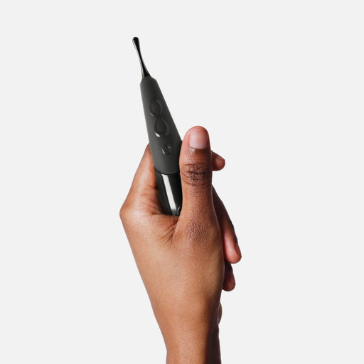 A hand holding the Zumio E - Rechargeable Pinpoint Clitoral Stimulator in black, reminiscent of the sleek design seen in Zumio products, is set against a white background. The stimulator features two buttons, a power symbol, and a small protruding tip for pinpoint stimulation.