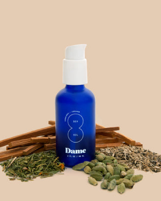 Dame Sex Oil for Intimate Massage blue bottle with ingredients surrounding bottle on beige background 