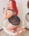A woman with vibrant red hair and tattoos smiles playfully while seated in a stylish white chair, holding a Sportsheets Saffron Classic Vegan Leather Flogger over her shoulder, exuding confidence and a touch of mischief.
